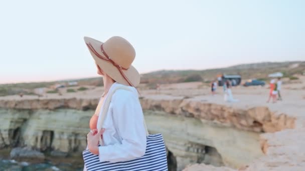 一个快乐的白人年轻女子站在悬崖边 头戴帽子 欣赏美丽的大海美景的背景画像 吸引人的女孩度假放松地看着大海玩乐 — 图库视频影像