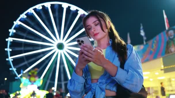 迷人的 可爱的 年轻的白人女子的画像 她站在游乐园里 晚上带着迷人的东西 手里拿着智能手机在网上交流 可爱的女孩在用电话聊天 — 图库视频影像