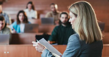 Beyaz kadın profesörün arkasından yaklaş gazetelerden bir ders oku ve üniversitedeki öğrencilere açıkla. Bilgi kavramı. Üniversitedeki öğretmenden ders çalış..