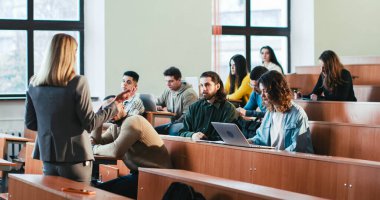 Kolejdeki kadın profesörün elinde kağıtlarla sınıftaki çok ırklı öğrencilere materyalleri anlatıyor ve anlatıyor. Kadın öğretmen konuşma yapıyor. Erkek öğrenci soru soruyor..