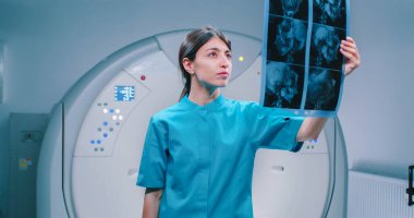 Mavi ceketli genç hemşire MRI taramasını dikkatlice inceliyor. Manyetik rezonans görüntülemesi için kapsülün arka planında çalışan bir kadın doktor. Muayene sonucu doktorda. Hemşire kameraya poz veriyor..