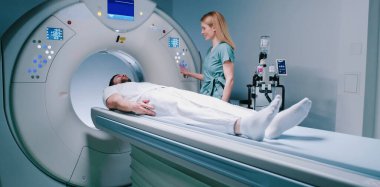 Erkek hasta TC tarama yatağında yatıyor. Adam MRI kapsülünden çıkıyor. Üniforma giymiş kadın doktor hastasıyla konuşuyor. Sağlık çalışanı gülümsüyor ve MRI kapsülünün düğmelerine basıyor.