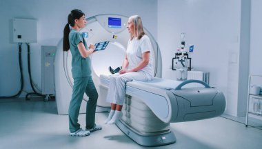 Doktor ve hasta tomografi sonuçlarını tartışıyorlar. Kadın doktor elinde tablet tutuyor ve beyin taramalarını yakınlaştırıyor. Kadın CT tarama yatağında oturuyor ve dikkatle doktoru dinliyor..