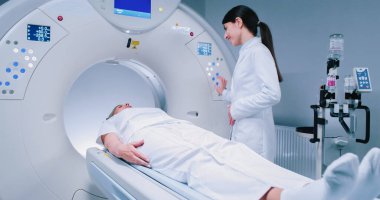 MRI ameliyatından önce önlük giymiş bir kadın doktor. Kadın doktor hastayı manyetik rezonans prosedürüne hazırlıyor. Hasta CT yatağında uzanıyor ve doktoru dinliyor..