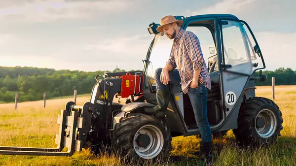 Close up of Caucasian handsome young male farmer in hat standing at tractor, smiling and looking at field. Countryside farming concept. Attractive man having rest from work at farm. Outside.