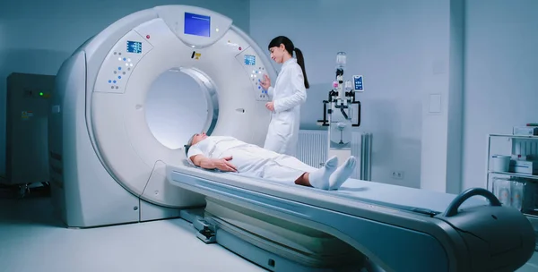 Senior woman in bathrobe undergoes MRI examination. Head doctor performs magnetic resonance imaging. Medical worker touches buttons and raises table with patient in MRI capsule.