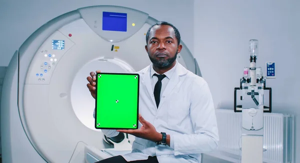 African American doctor is posing for camera. Mature man is showing screen of chroma key tablet. Multicultural doctor is smiling and looking at camera. Medical worker at background of MRI capsule.