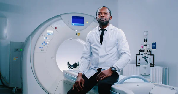 African American doctor at room of magnetic resonance tomography. Shoot of male medical worker posing at camera. Multicultural man put hands on knees and looking straight at camera.