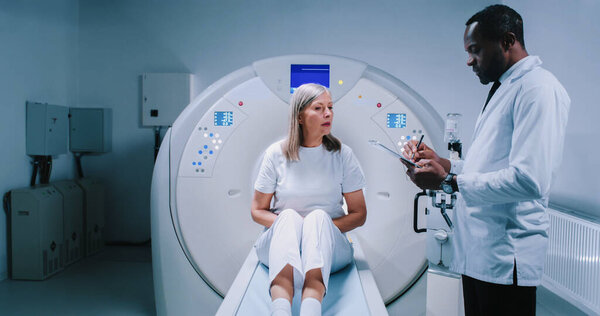 Врач и пациент обсуждают это в современной томографической лаборатории. Афроамериканский врач держит папку и жесты. Женщина сидит на кровати МРТ и готовится к магнитно-резонансной томографии..