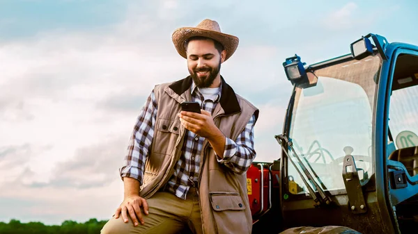 Çiftlikteki Neşeli Genç Erkek Sürücü Traktörün Önünde Duruyor Akıllı Telefondan - Stok İmaj
