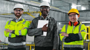 İnşaat alanındaki üç adam kameraya bakıyor ve gülümsüyor. Baş mühendis dizüstü bilgisayar tutuyor. Üniformalı iki çok kültürlü işçi kollarını kavuşturdu. Etnik çeşitlilik.