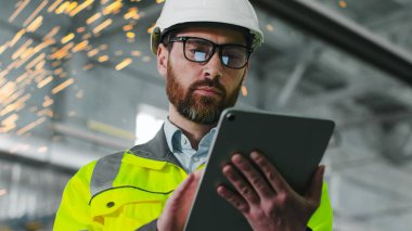 İnşaat alanında olgun sakallı bir adam. Üniformalı ve gözlüklü mühendis iş akışının tablet geçmişini tutuyor. Çalışanlardan biri atölyesindeki aletine bir şey not etmiş. Proffesyonların çeşitliliği.