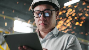 Kameraya bakıp tableti tutan Asyalı erkek mühendisin portresi. Beyaz gömlekli ve çarşaf giymiş bir işçi. Kaynak işinde kıvılcımlar saçıyor. Gözlüklü eleman şantiyede çalışıyor..