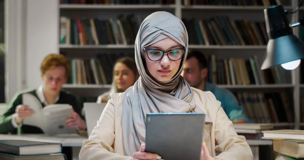 Young Arab muslim woman in hijab and glasses working on tablet in library and scrolling on screen. Arabian beautiful female student in headscarf holding computer. Tapping on gadget. Education concept.