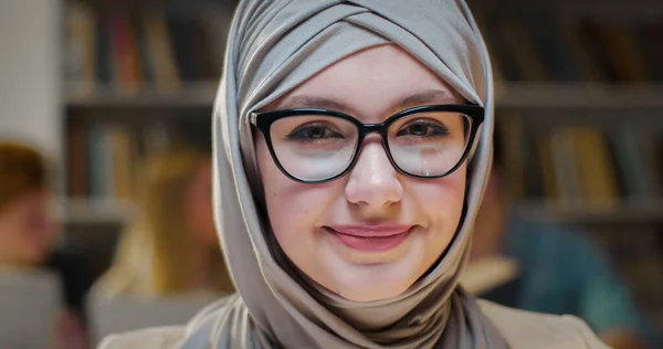 Close up of beautiful young Arab muslim woman face in hijab and glasses sitting in library space and looking with smile to camera. Pretty female student in Arabian headscarf in bibliotheca. Portrait.