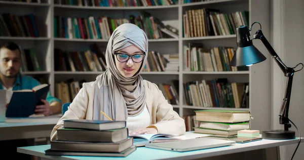 Young muslim female student in hijab and glasses sitting at table in library, reading book and making marks on pages. Arab woman in Arabian headscarf studying with textbook and marking.