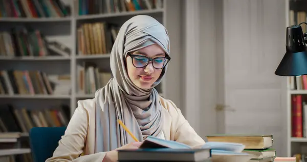 Young muslim female student in hijab and glasses sitting at table in library, taking notes and reading textbook. Woman in arabian headscarf noting and learning from book. Bibliotheca study concept.