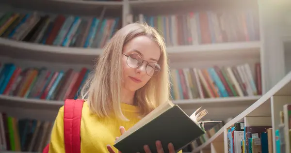 Rear of Caucasian young blonde beautiful woman in glasses walking in library passage, then choosing texbook from shelf and reading it. Female student flipping pages of book. Education concept.