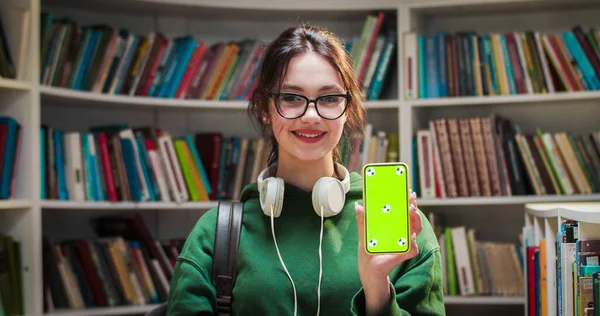 Portrait of young Caucasian pretty female student in glasses smiling and showing smartphone with green screen and tracking motion in library. Chroma key. Woman with mobile phone demonstrating.