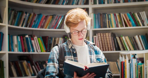 Caucasian red-haired male student in glasses and headphones flipping pages, reading book and searching for information in library. Guy with red hair with textbook at bookshelves listening to music.