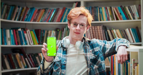 Portrait of young Caucasian red-haired male student in glasses smiling and showing smartphone with green screen and tracking motion in library. Chroma key. Guy with mobile phone demonstrating.