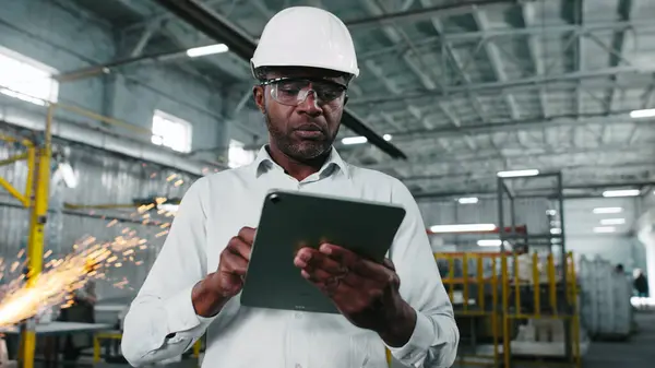 African American engineer in hard hat is looking through workshop. Man is holding gadget at construction site. Worker makes notes in tablet. Employee on background of welding sparks flying.