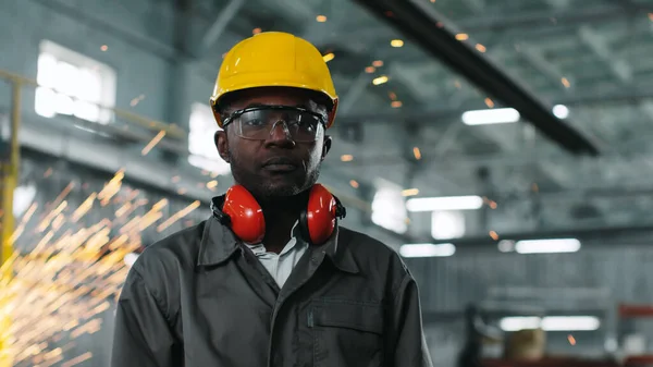 African-American worker wearing hard hat and goggles is staring intently at camera. Employee looks calm on working background of production. Ethnic diversity in construction.