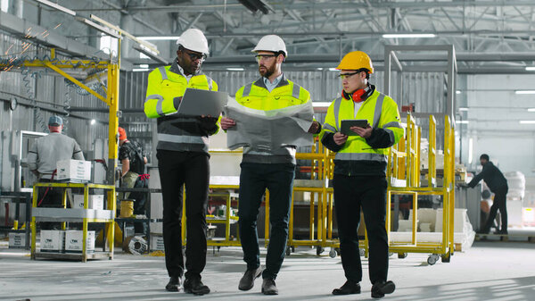 Tree construction workers. Men in special uniforms and helmets move in the workshop. Engineers in full growth in production. Diversity of employees. Workers discuss project with laptop and blueprint.