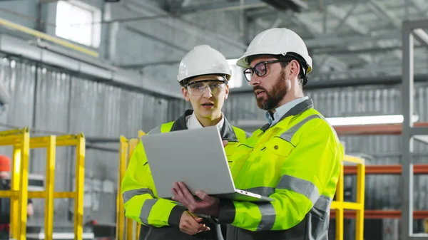 Main engineer briskly explains plan of action to employee. Bearded man holding notebook and gesturing. Workers in yellow jackets and helmets cooperate at construction site.
