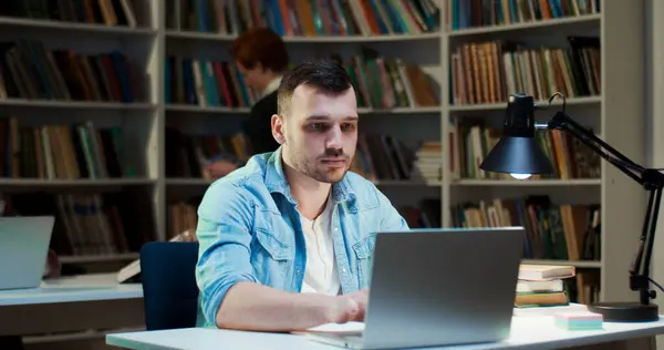 在图书馆的笔记本电脑前学习的白人年轻人 男人在桌上做笔记 用铅笔在笔记本上写字的人 在书目中工作A 学习和工作概念 搜寻资料 图库图片