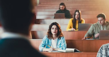 Young pretty Caucasian female student listening lection at college, writing down and using textbook. Reading book at seminar at high school. University study concept. Studying in classroom. clipart