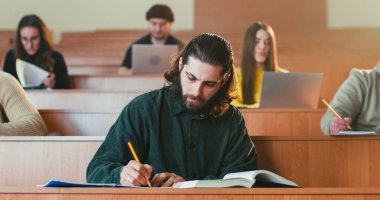 Oditoryumda oturup ders kitabı okuyan, notlar alan ve kameraya gülümseyen yakışıklı beyaz erkek öğrencinin portresi. Adam kitap okuyor ve derste ilginç bir şey işaretliyor. Çalış.