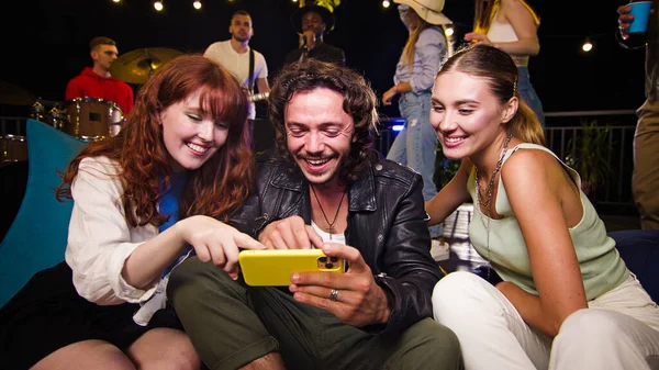 Company of young friends watching funny videos on a telephone. Rooftop party friends having fun listening to band performing at a concert on party.