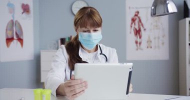Dolabın masasında otururken çalışan ve cihazı tıkırdatan oldukça mutlu bir doktor. Beyaz önlüklü ve tıbbi maskeli güzel beyaz kadın doktor hastane ofisinde tablet kullanıyor. Sağlık kavramı