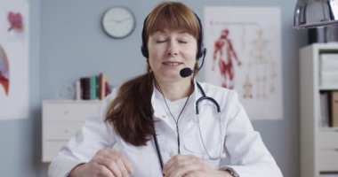 Beyaz önlüklü ve kulaklıklı güzel beyaz bir doktor ofisinde oturmuş kamerayla konuşuyor. İnternette sağlık kuralları öğreten mutlu bir kadın doktor blogcusu. Tıbbi video günlüğü kavramı