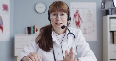 Beyaz önlüklü, gözlüklü, ofisinde oturan ve kameraya konuşan güzel beyaz bir doktor. Kulaklıklı neşeli kadın doktor blogcu internette sağlık tavsiyesi veriyor. Tıbbi video günlüğü kavramı
