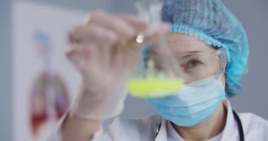 Maskeli, gözlüklü ve eldivenli beyaz kadın epidemiyoloji uzmanının ellerinde Coronavirus 'a karşı sıvı aşıyla cam matara tutmasını istiyorum. Aşıların laboratuvar testleri. Klinik araştırma laboratuarı kavramı