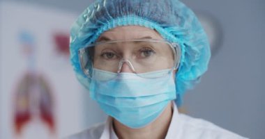 Ofisteki kameraya bakan, tıbbi maskeli ve gözlüklü güzel beyaz kadın epidemiyolojistin portresini yakından çek. Hastane ofisindeki güzel laboratuvar asistanı. Sıhhiye kavramı