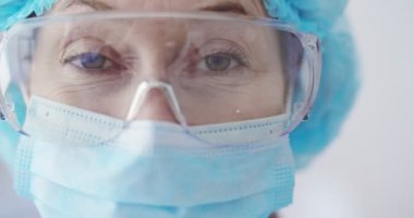 Tıbbi maskeli ve gözlüklü kameraya bakan kadın epidemiyolojistin yakın portresi. İçerideki hastane ofisinde kadın laboratuvar asistanı. Tam koruma kostümü giymiş bir sağlık görevlisi. Laboratuar kavramı