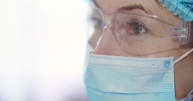 Kafkas kadın epidemiyolojistin tıbbi maskeli ve gözlüklü portresini yakından çek ve kameraya dön. Tam korumalı kostüm giymiş laboratuvar asistanı içerideki kameraya bakıyor..