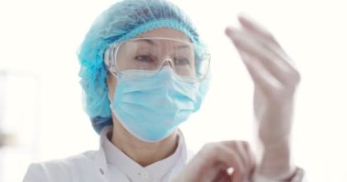 Tıbbi maskeli, eldivenli ve gözlüklü, odada kameraya bakan beyaz kadın epidemiyolojistin yakın portresi. İçeride tam koruma kostümü giymiş laboratuvar asistanı. Laboratuvar kavramı.
