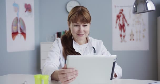 漂亮而快乐的白种人女医生穿着白色长袍坐在医院的办公室里用平板电脑 非常快乐的医生在橱柜的桌子边工作和敲敲设备 医疗概念 — 图库视频影像