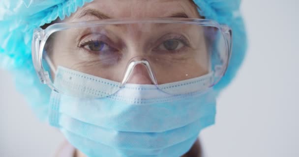 用医疗面罩和护目镜近距离拍摄白种人女性流行病学家的肖像 看着办公室里的摄像机 医院办公室室内漂亮的实验室助理 身穿全套防护服的医务工作者 — 图库视频影像