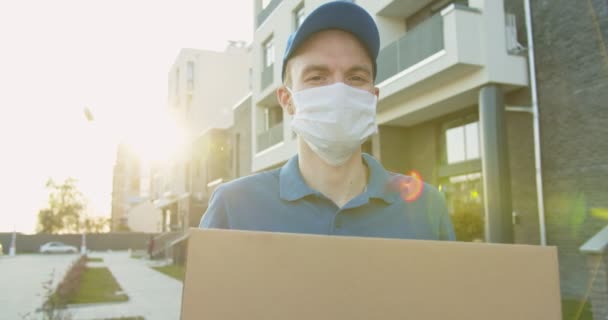 一个配送工人头戴蓝色帽子 戴着医疗面罩 手拿着纸盒在街上走着的画像 检疫期间运送纸箱包裹的快递员 待在家里 上网订购 — 图库视频影像