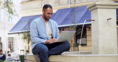 Şehir merkezindeki çeşmede dizüstü bilgisayarı ve klavyesinde daktiloyla oturan gözlüklü yakışıklı bir genç. Açık havada. Şehirde bilgisayar üzerinde çalışan gözlüklü serbest çalışan yakışıklı bir erkek..
