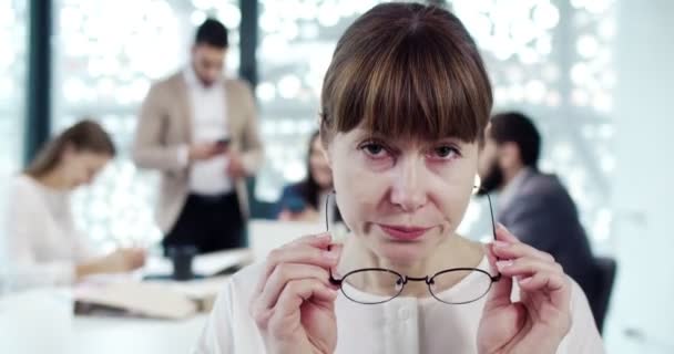 カメラを見て眼鏡をかけている有名な会社のオフィスのマネージャーとして働く成熟した厳格な女性の肖像画 バックグラウンドの労働者を持つ深刻な慎重な白人女性の従業員 — ストック動画