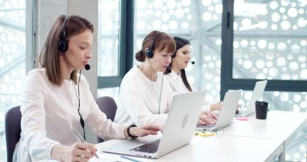 クライアントと話すためにラップトップとヘッドフォンを使用して現代コールセンターオフィスの3人のプロフェッショナルな効果的な女性労働者の側面図 職場で美しいカスタマーサポート担当者 — ストック動画