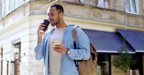 スマートフォンでコーヒーを飲んでテキストメッセージを送る 素敵な日に立っているメガネの若いハンサムな男 アウトドア 笑顔の男性旅行者 町でチャット 携帯電話で飲み物を飲みに行く — ストック動画