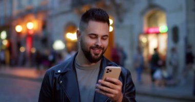 Siyah deri ceketli neşeli genç adam şehir caddesinde duruyor ve akıllı telefondan mesaj atıyor. Açık havada. Kasaba meydanında yakışıklı bir erkek. Sohbet ediyor, gülümsüyor ve telefonla konuşuyor..