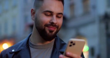 Deri ceketli, gülümseyen, cep telefonuna mesaj atan ve mesaj okuyan neşeli, genç ve yakışıklı bir erkek. Dışarı çıkın. Sakallı çekici bir adam akıllı telefondan tıklıyor ve kaydırıyor..